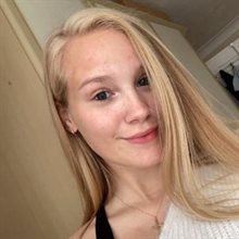 Head and shoulders photograph of a young white woman with long blonde hair wearing a white jumper over a black top standing in front of a wardrobe