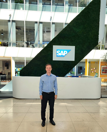 Matthew Thompson smiling wearing a blue shirt and black trousers standing in front of the SAP logo in a large foyer with glass windows and a grey tiled floor