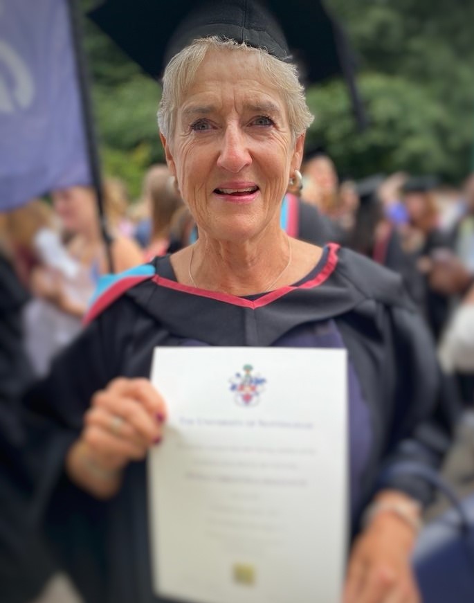 Fiona on graduation day, wearing a black motarboard hat and gown, holding her degree certificate