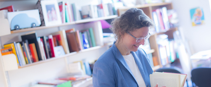 Academic stands in her study wearing a blue jacket in front of a bookcase leafing through a book