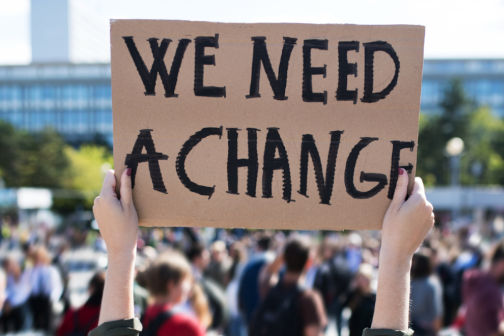 Two hands holding up a placard at a demo saying "We need a change"
