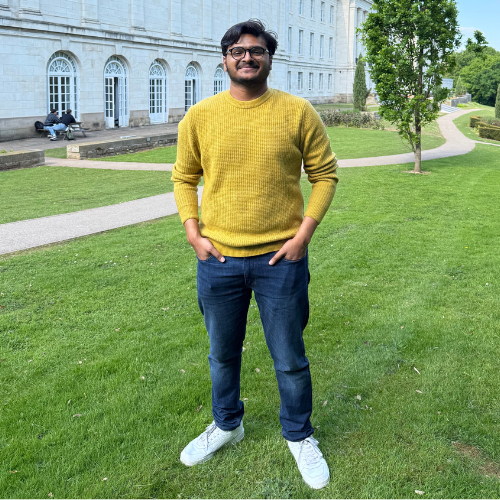 Aakash Parameswaran with black short curly hair wearing a bright yellow jumper, grey trousers and white trainers standing on grass in front of white Trent Buiding at the University Park Campus in Nottingham