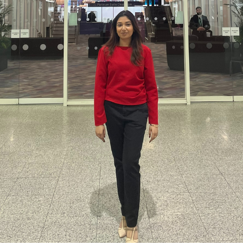 Aiswarya Krishnan with long black hair, wearing a red jumper and black trousers standing in front of large glass doors below a bright Enactus sign