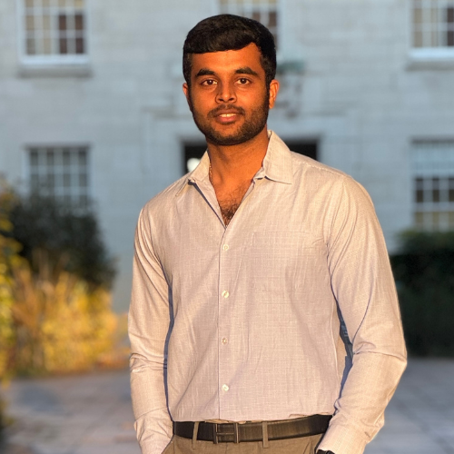 Anudeep Sridhar, with short black hair and a beard, wearing a white shirt, stands in front of a large grey stone building.