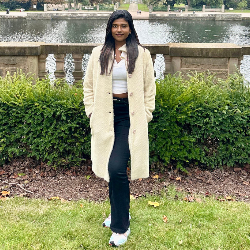 Clara Benita Jesula Justin Thomas with long black hair wearing a long cream coat and white top, black trousers and white trainers standing on grass in front of the lake at the University Park Campus in Nottingham