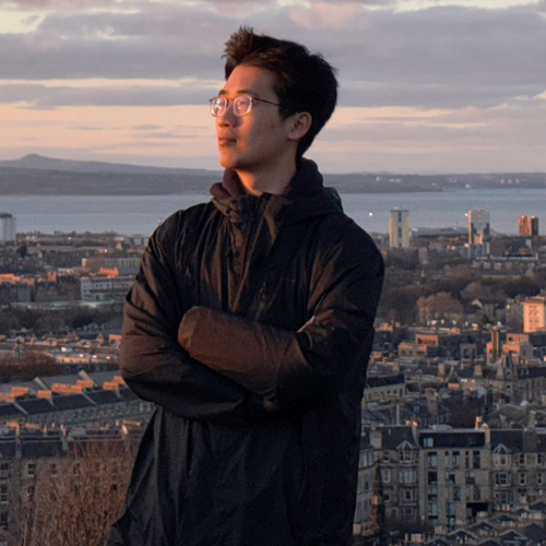 Hongda Jin wearing a black coat with arms folded standing on a hill in front of a city with pink clouds at sunset
