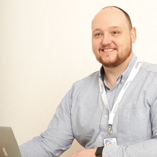 Photo of Lloyd Major. Lloyd has very short hair and is sat at a table with a laptop open. he is wearing a grey shirt with a lanyard around his neck.