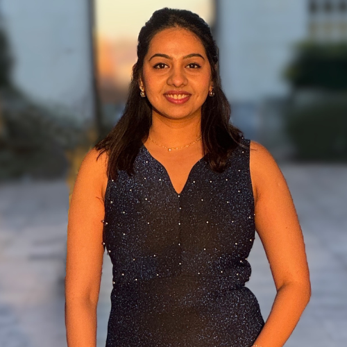 Shreya Dathathraya with long black wavy hair smiling, wearing a black glitter dress and silver shoes, standing in front of the white brick Trent Building at the University Park campus