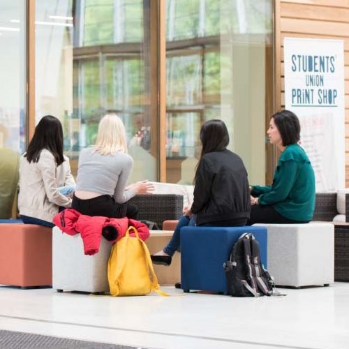 Group of students sat in the Exchange building with a sign in the background saying 'Student Union Print Shop'