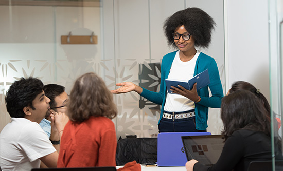Female PGR student standing talking to other PGR students who are sitting round a table