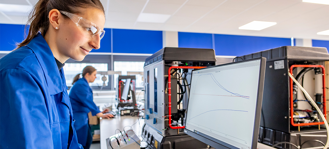 Students in blue overalls viewing data charts on computer screens in a geotechnics and fluid mechanics lab with machine equipment visible on the desks