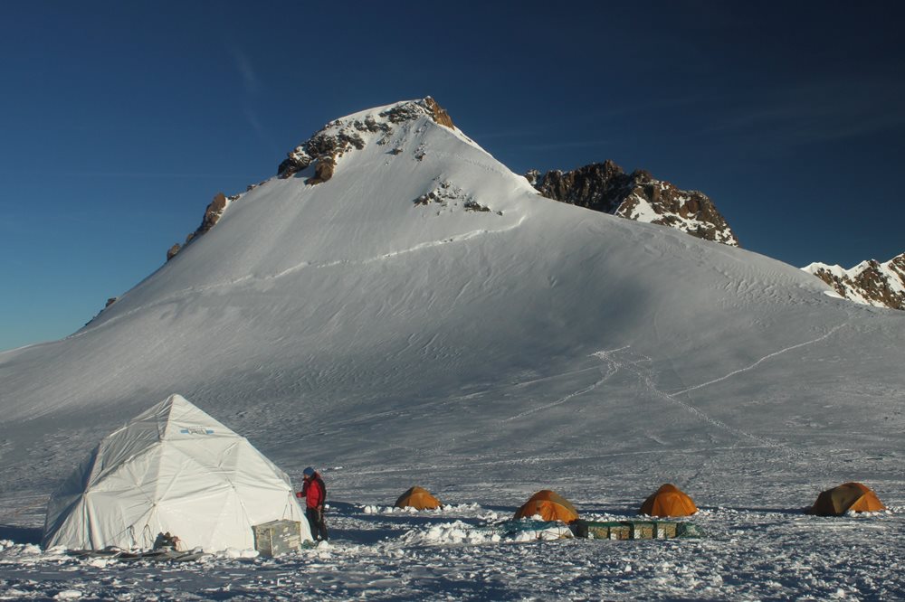 At the base of a snowy mountain are four orange tents and one white dome tent.
