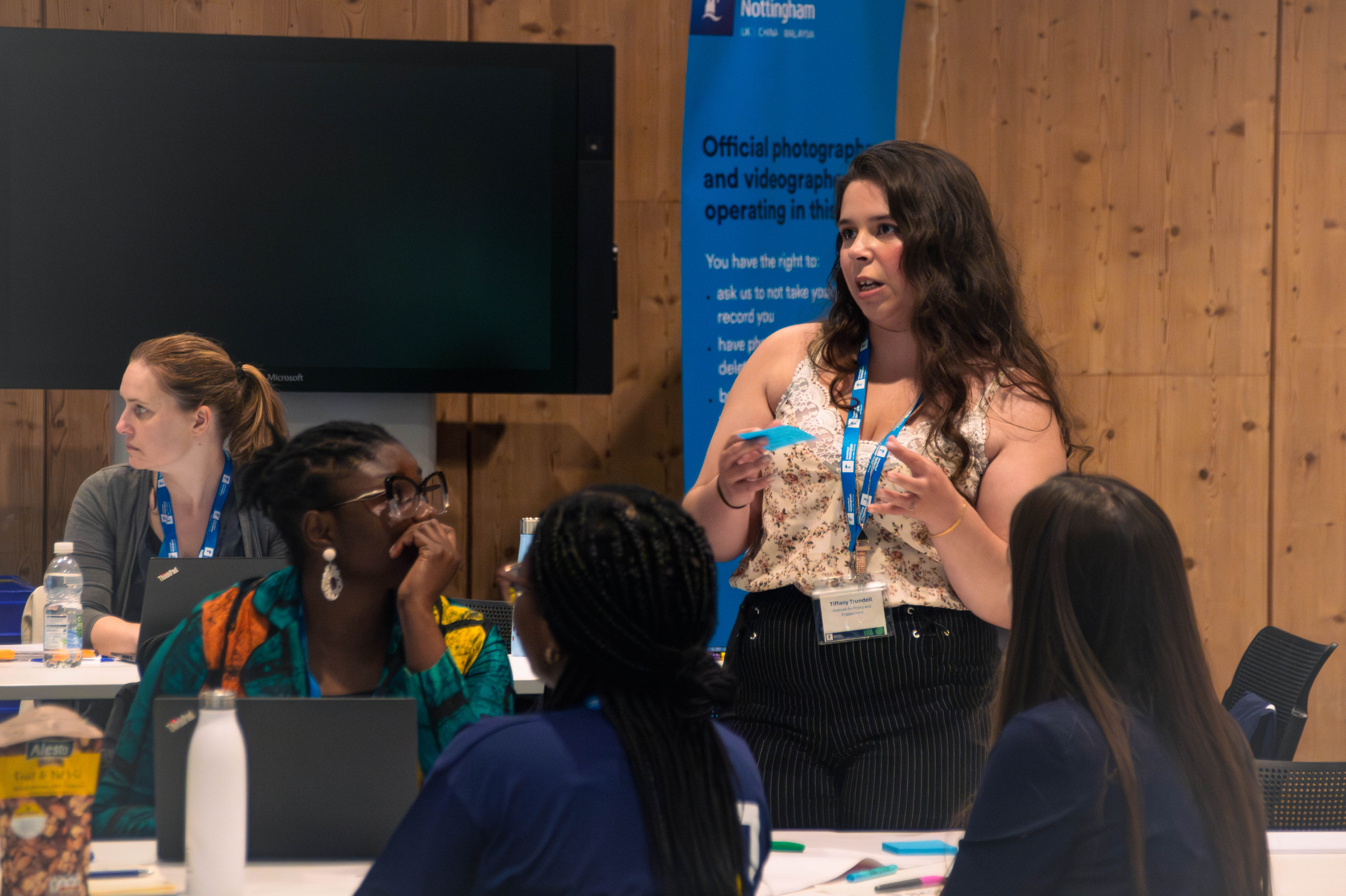 A white woman standing and talking to the audience. She is sitting with a group at the table