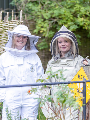 Two people standing in an outdoor setting, one of them is wearing a beekeepers outfit
